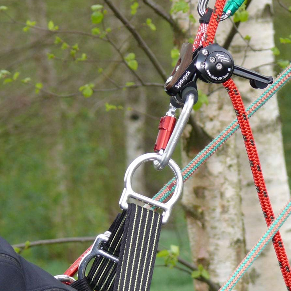 Close-up of a climbing harness and ART Spiderjack 2.1 Dyneema, in use with red and green ropes, a black harness strap, and metal hardware against a blurred tree background.